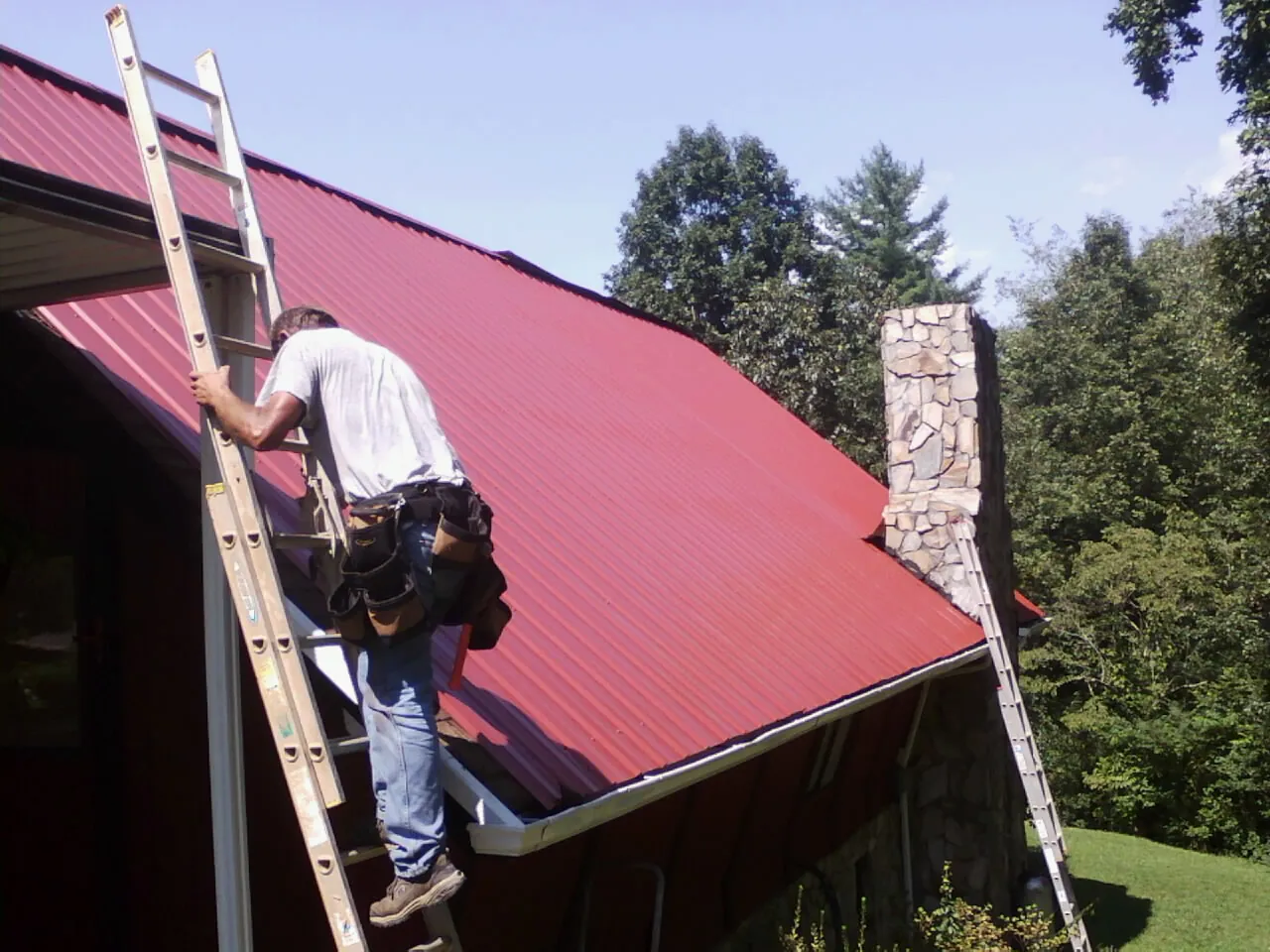 Roofer on a ladder beside a red metal roof and stone chimney — Soffit & Fascia Repair by Roofing Brothers in Grand Lake